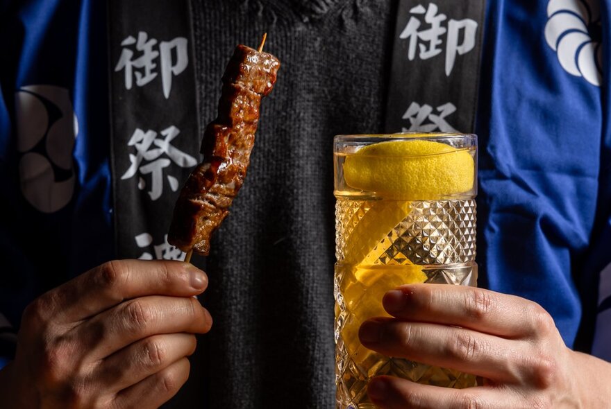 A person holding a drink in a tall glass in one hand and a meat skewer in the other hand, wearing a blue and black cotton gown inscribed with Japanese characters on its lapel.