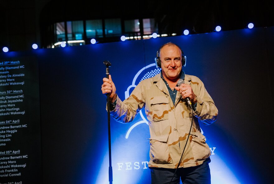 Comedian Jimeoin on stage with headphones holding a microphone stand at the Arbory Bar's Silent Comedy Festival.