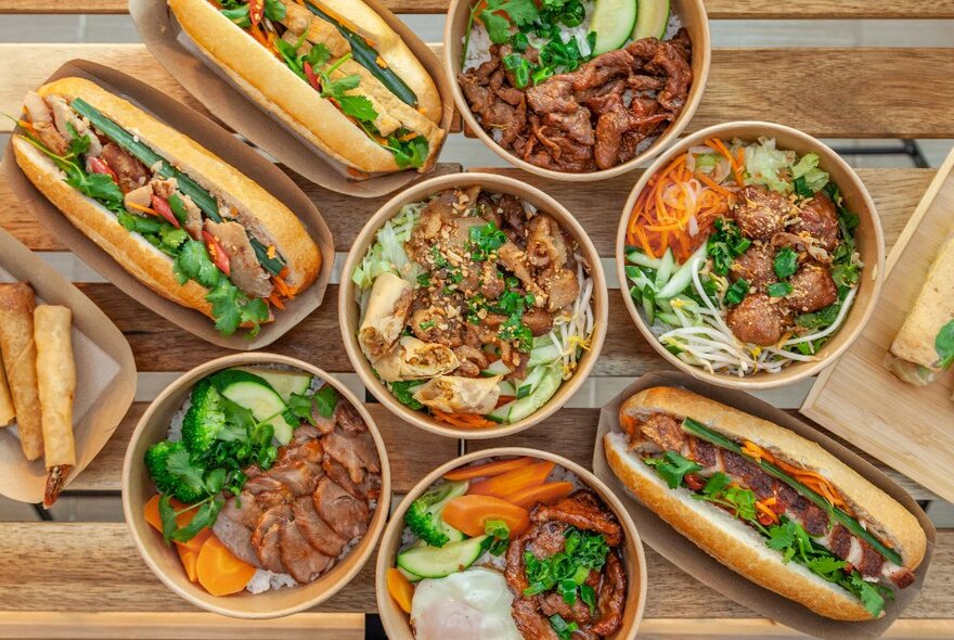 Bowls of Vietnamese dishes and filled banh mi baguettes on a wooden table.