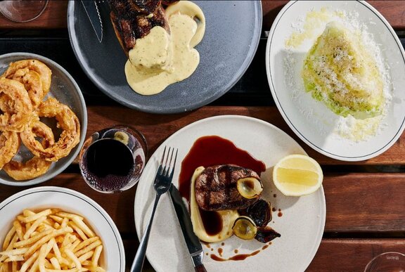 A steak on a plate covered in a sauce, with fries and a salad  on side plates.