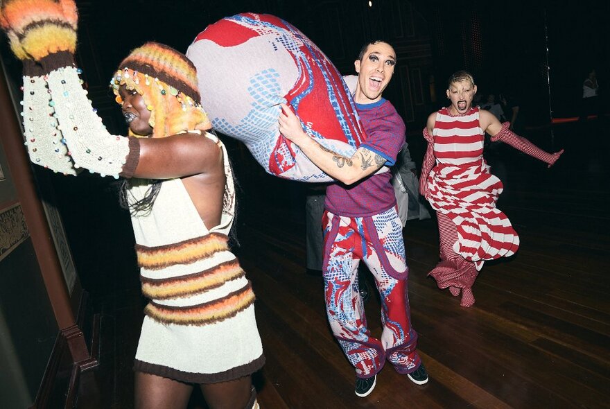 Three fashion models in bright outfits smiling in a darkened space,  backstage at a fashion show.