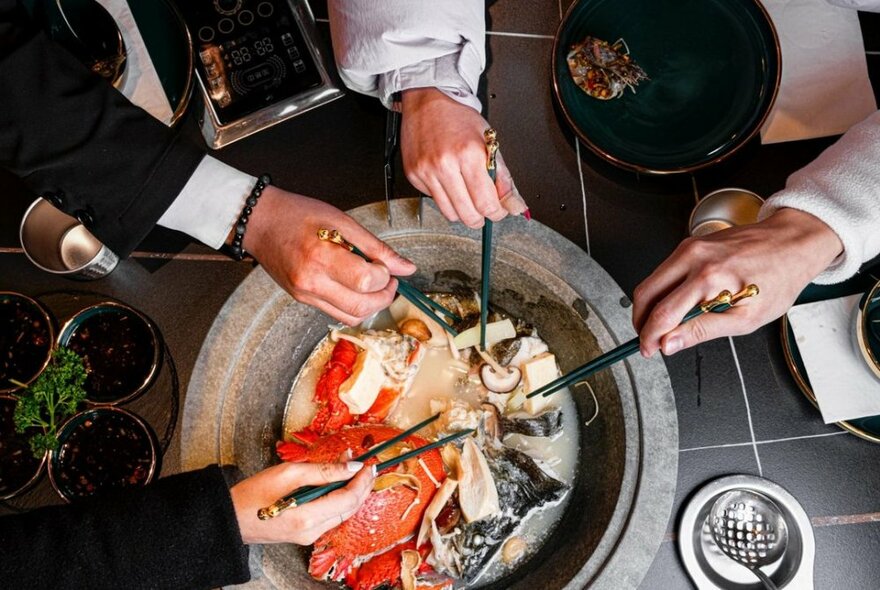 Hands holding chopticks over a central stone cooking pot containing fish.