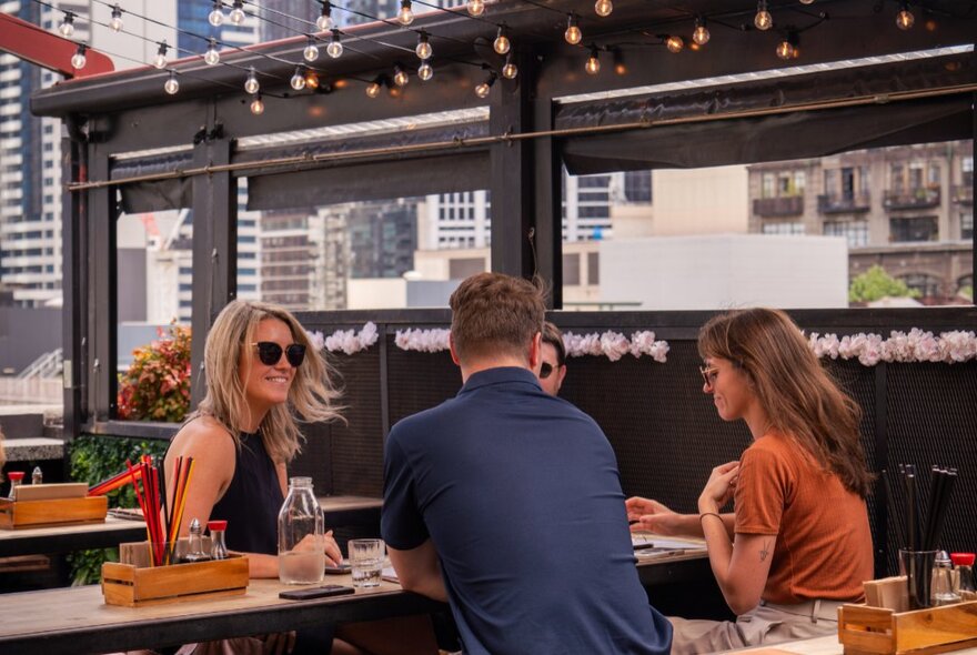 A table of friends enjoying a drink at an outdoor rooftop bar, with views of Melbourne city buildings in the background.