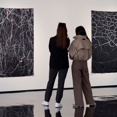 Two women in a gallery with white walls and highly polished black floors observing two pieces of abstract photographic art hung on the wall.