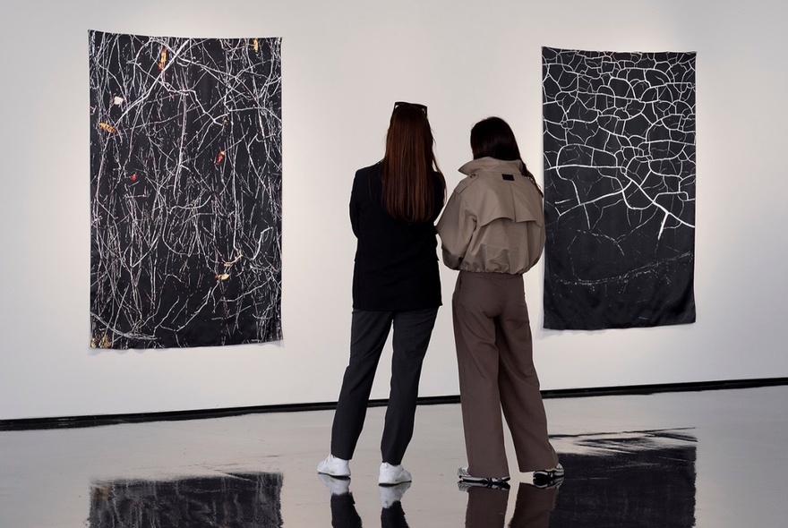 Two women in a gallery with white walls and highly polished black floors observing two pieces of abstract photographic art hung on the wall.