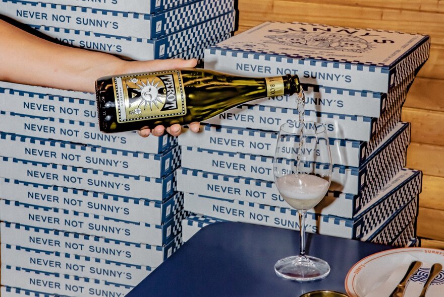 Hand pouring sparkling wine into a glass, stacks of pizza boxes in the background.