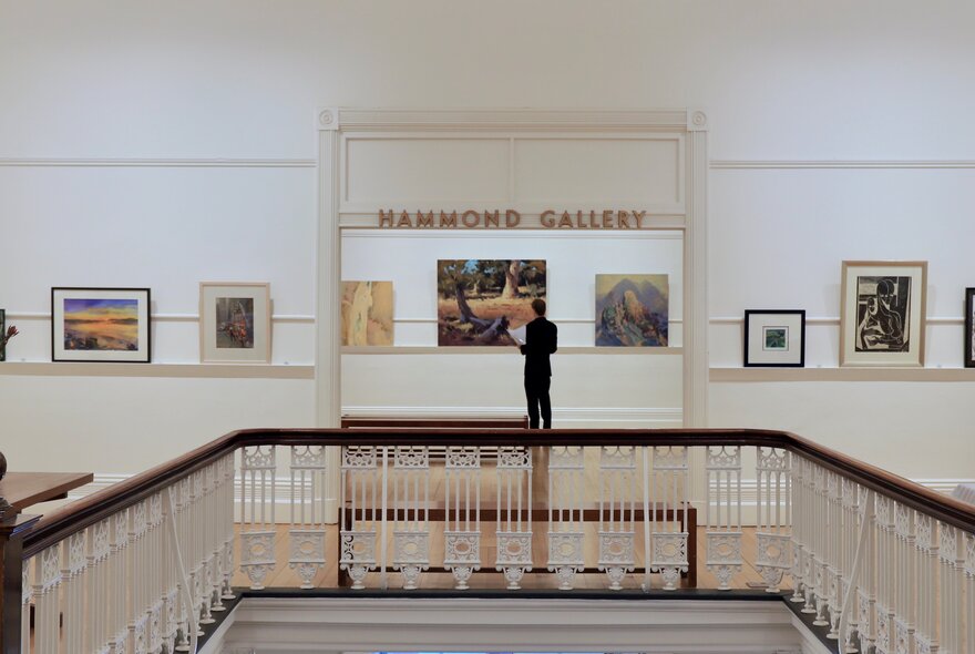 A gallery space with works on white walls behind a lacework staircase balcony.