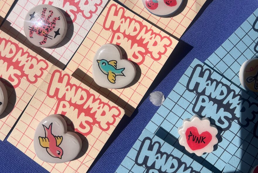 Handmade pin badges and enamel pins displayed on grid-patterned backing cards labelled 'Handmade Pins'.