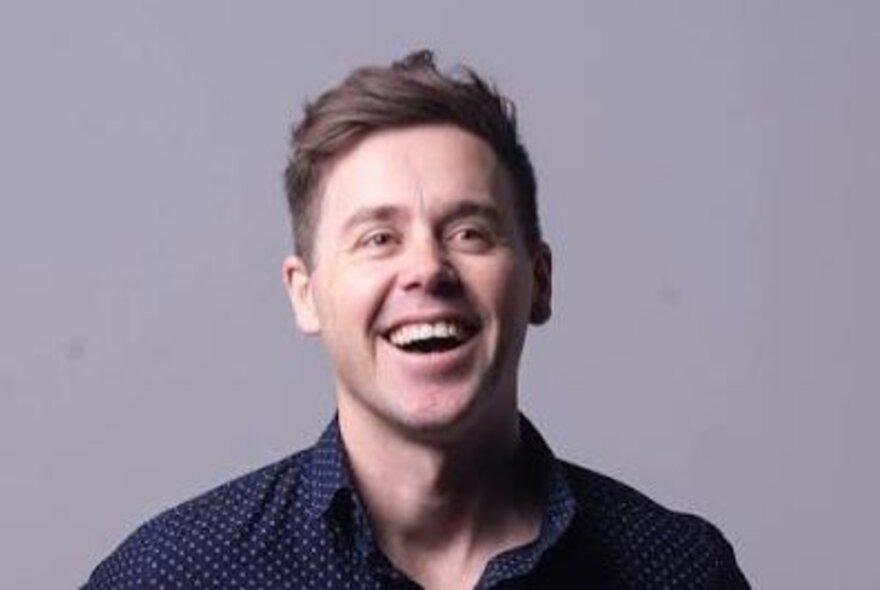 Comedian Josh Earl laughing, he is a middle aged man wearing a blue shirt posed against a light grey background.