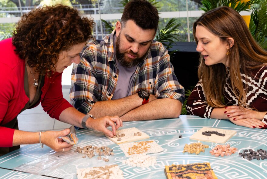 Three people with heads together looking at a tiny piece of mosaic at a craft event. 