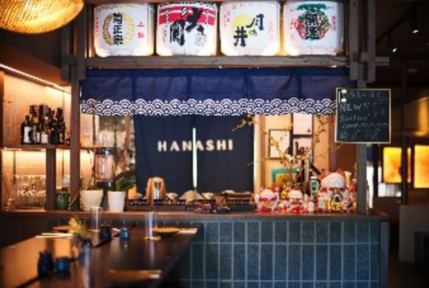 Interior of Hanshi restaurant showing a central bar area and high dining tables and stools, decorative lanterns and sake barrels on display.