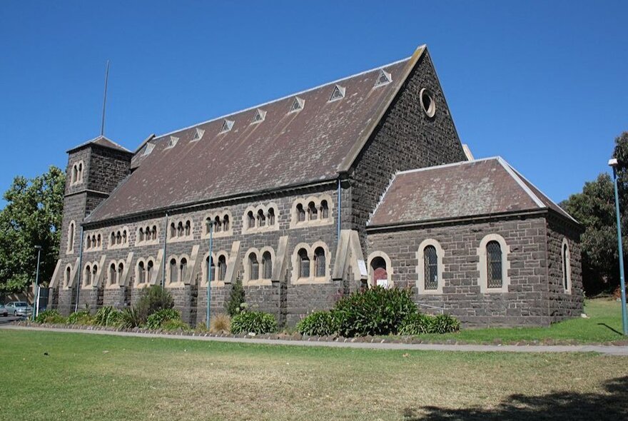The bluestone exterior of the Church of all Nations in Carlton, surrounded by green lawns, set against a clear blue sky.