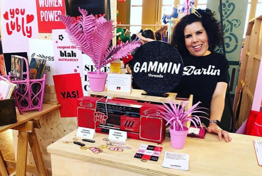 A smiling woman wearing a black t-shirt with the word Darlin printed on it in white letters, standing behind a small table of Gammin Threads material and products on display.