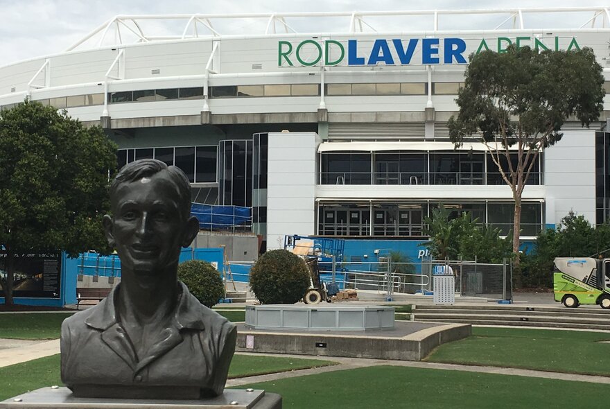 The exterior of the tennis stadium Rod Laver Arena at Melbourne Park, with green lawns and a bronze bust of Rod Laver in the foreground.
