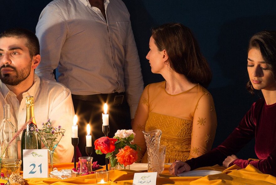 Three people seated at a banquet table, with a fourth person standing behind them. Each person faces a different direction. The table is set with burning candles, empty bottles, wilting flowers, and Arabic-language place cards.