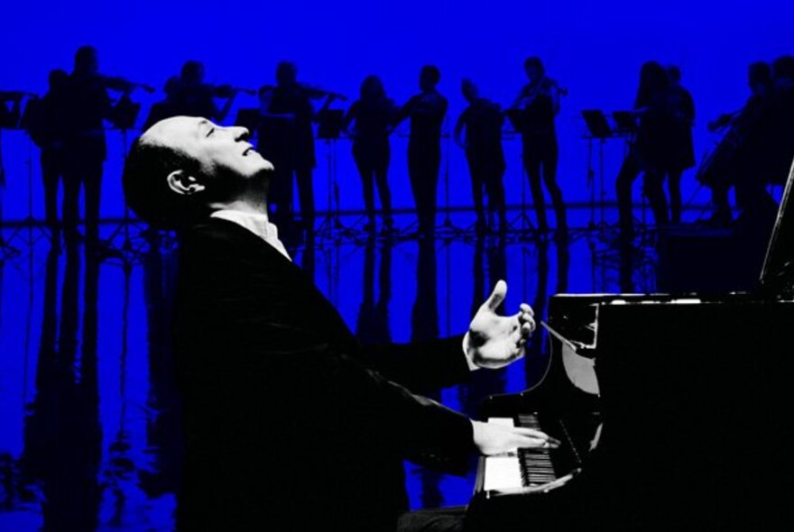 A man seated at a piano and playing it with his head slightly tilted back, in the background an ensemble of standing orchestra musicians are silhouetted against a deep blue background.