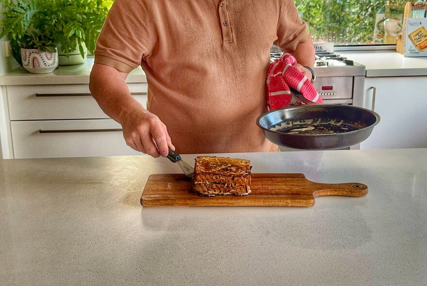 Person placing a toastie from a frying pan onto a wooden board in a kitchen.