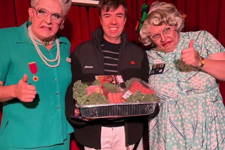 Two men dressed as elderly ladies wearing wigs, dresses and jewels with an audience member who is holding a meat tray, on stage at Granny Bingo.