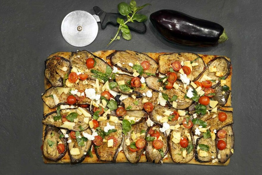 Large rectangular pizza next to pizza cutter and eggplant.