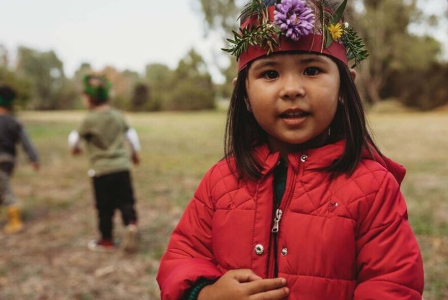 Child wearing a red puffer jacket and a floral headdress in a park setting.
