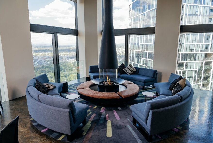 Circular couches arranged around a small round table and central fireplace at the Sofitel on Collins, with views of Melbourne city and surrounds though the large floor to ceiling windows. 