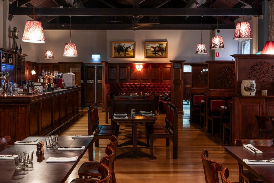 Interior of a restaurant showing wood panelled floors and seating booths, soft lighting, a central bar and paintings hanging on the rear wall.