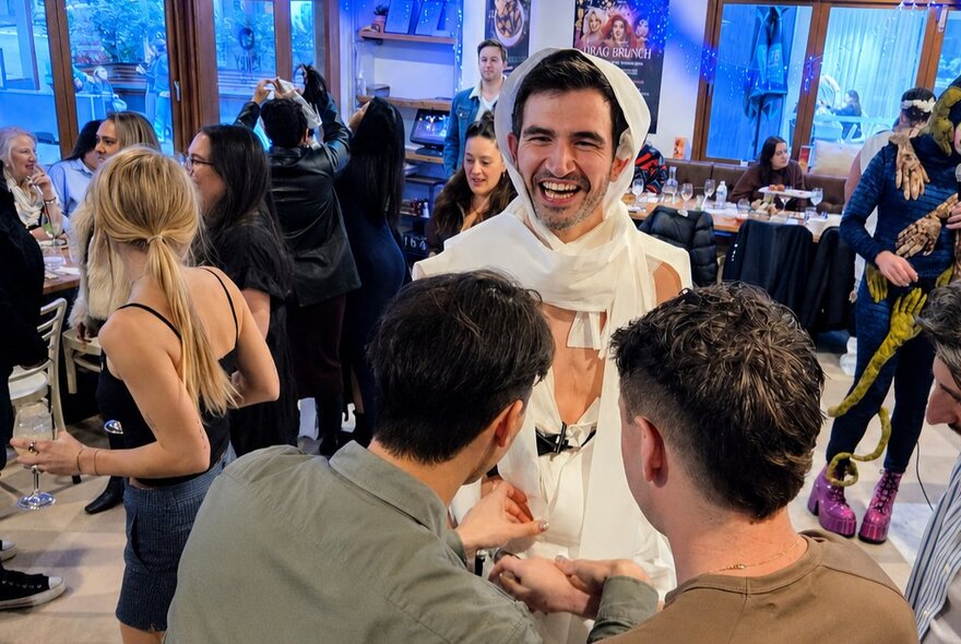 A man in a white hooded outfit made from toilet paper, laughing as he talks to two other men in a restaurant setting.