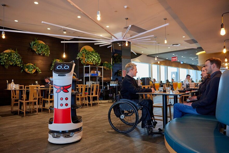 A group of people dining in a large restaurant. One is using a wheelchair. A robot waiter with cat features is driving past.