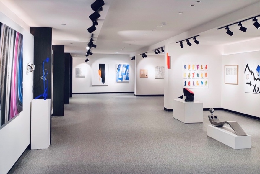 A gallery space featuring colouful artworks on the white walls and white pedestals.