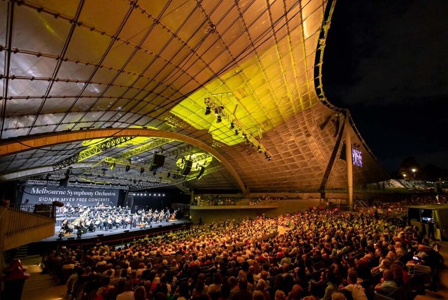 The Sidney Myer Music Bowl at night, with an orchestra playing on the stage and the rows of seats facing the stage filled with people.