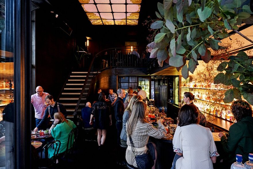 People seated at tables and on stools in a cocktail bar with illuminated shelves and stairs in the background.