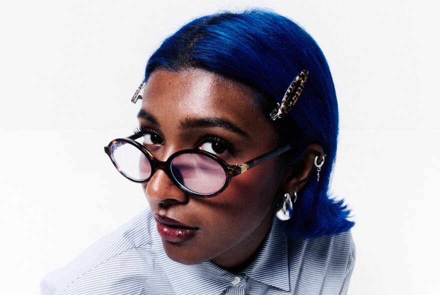 Singer Charlotte Dirckze posing for the camera, wearing glasses with her short blue hair slicked back. 