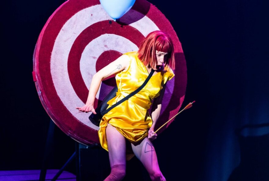 A performer in Circus Oz leaning against a target with an arrow in her hand and balloon near her head