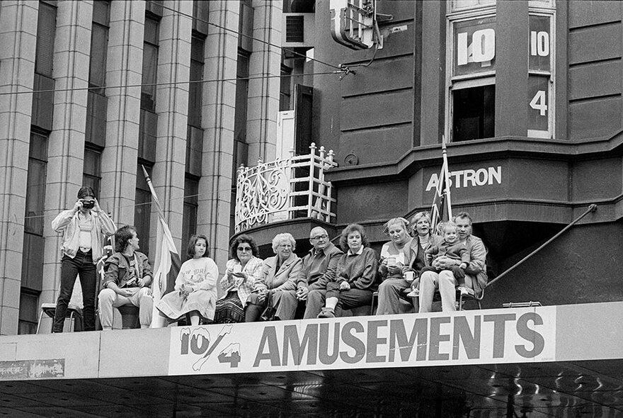 An old black and white image of people sitting on top of city  building verandahs watching something below.