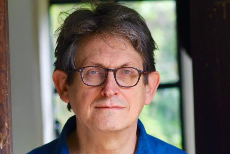 Journalist Alan Rusbridger wearing glasses and a blue shirt standing in front of a window with trees behind.