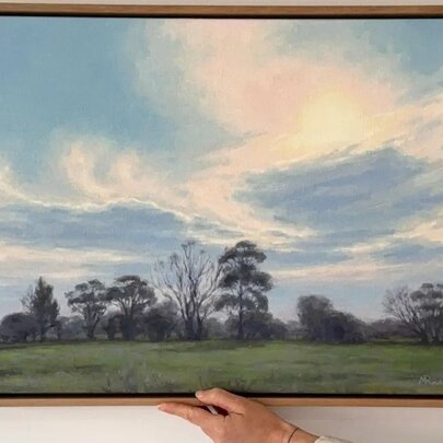 A person's hands holding up a timber-framed oil painting of a farmland landscape with blue skies.