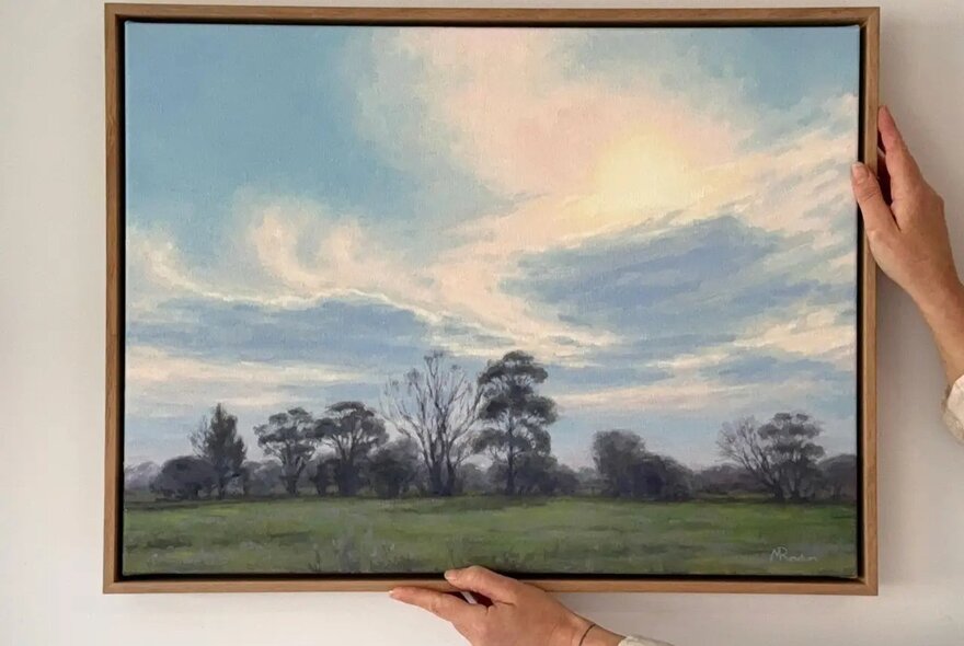 A person's hands holding up a timber-framed oil painting of a farmland landscape with blue skies.