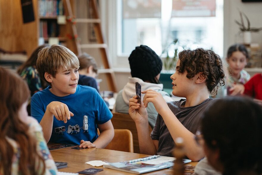 Children seated around work tables in a large studio space, interacting with one another.