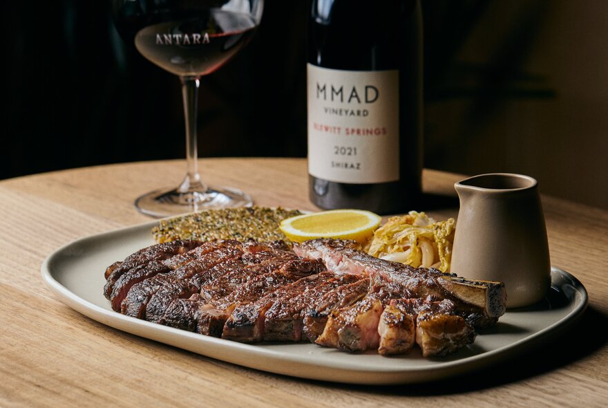 A rectangular plate with a long piece of steak sliced into thin strips, a small jug of sauce and a glass and bottle of red in the background. 