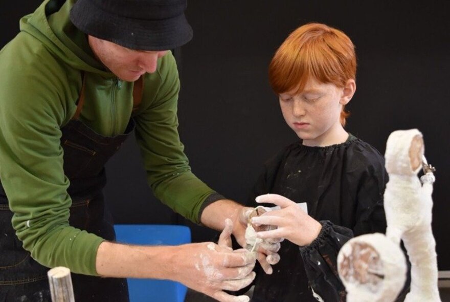 Adult showing a young boy with red hair how to sculpt a figure with clay.
