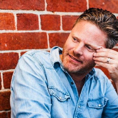 A man leaning his head against his hand as he sits smiling lightly against a red brick wall.