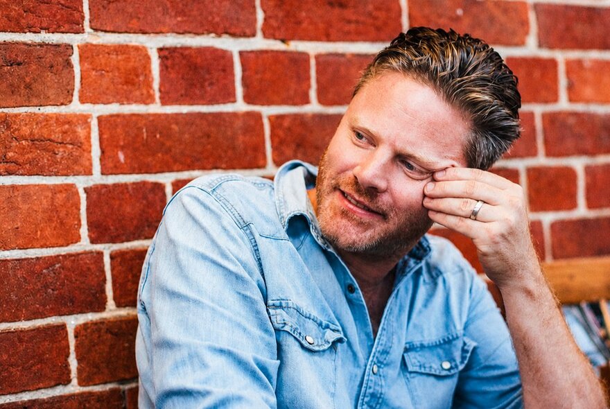 A man leaning his head against his hand as he sits smiling lightly against a red brick wall.
