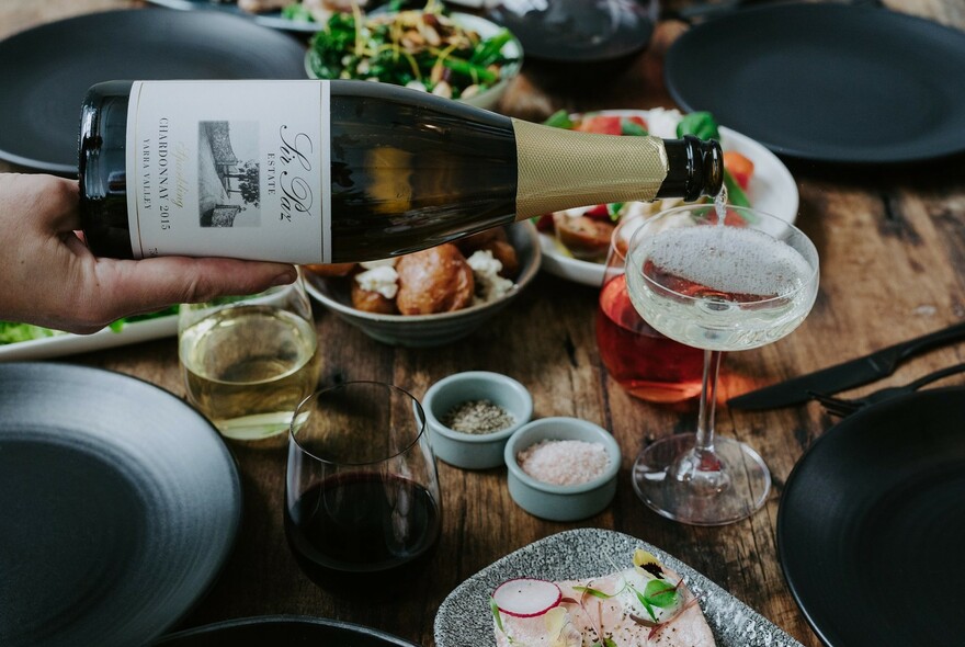 Dining table with champagne being poured into a wine glass alongside a selection of salads, meats and condiments.