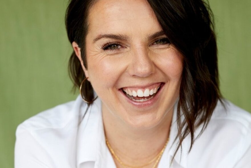 Kate Mildenhall, a woman smiling with an open mouth, she has brown hair, and is wearing a white shirt and posed against a pale green background. 