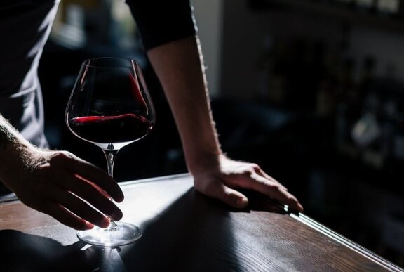 A person leaning over a table holding a large red wine glass.