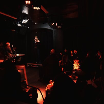Intimate, very dimly lit indoor club, with a stand-up comedy performer on stage and low-level amber and red stage lighting.