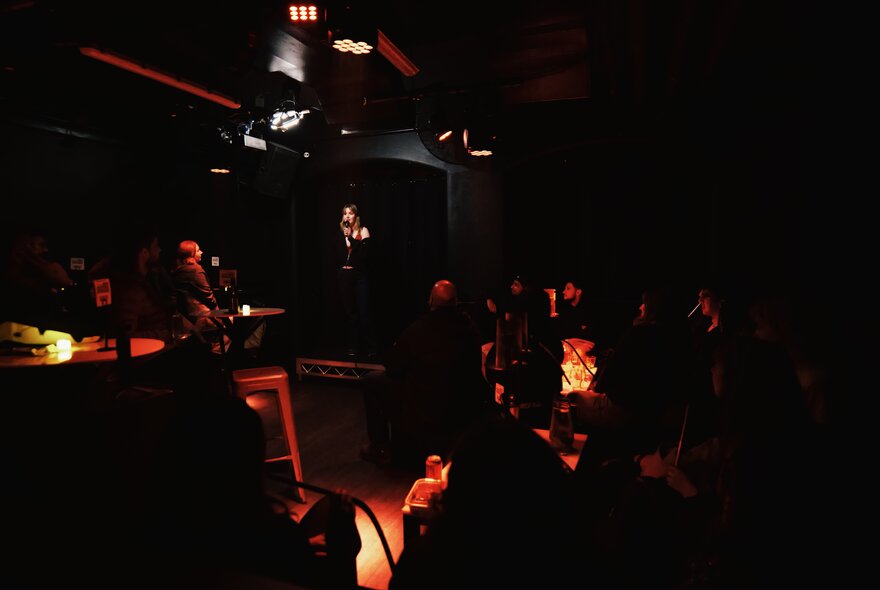Intimate, very dimly lit indoor club, with a stand-up comedy performer on stage and low-level amber and red stage lighting.