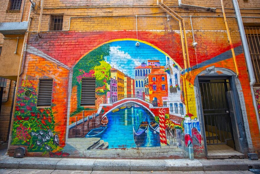 A mural of a European canal with colourful houses, flowers and a boat going under a bridge.