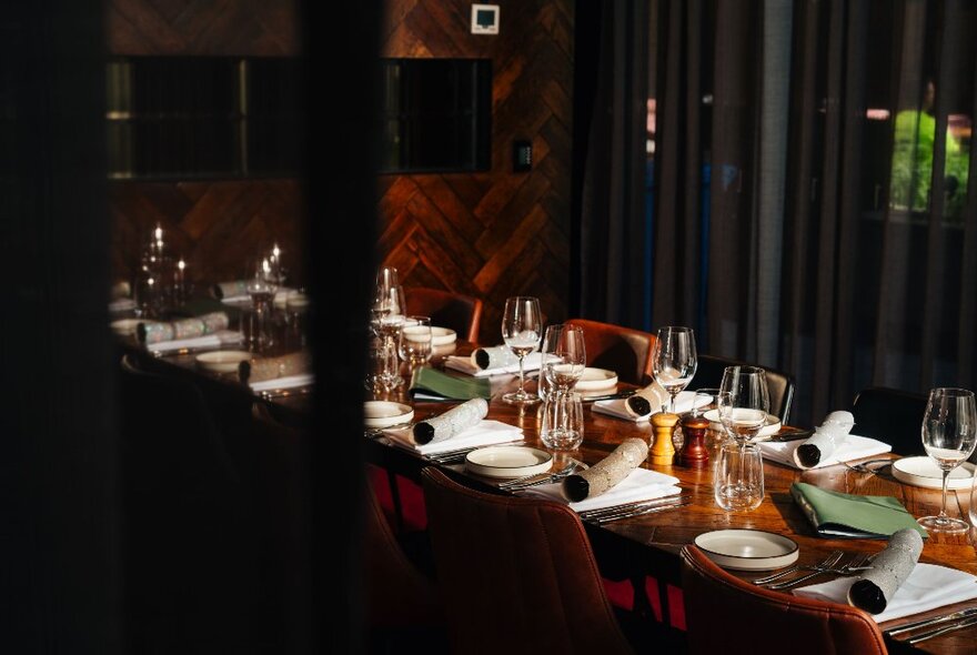 A long table set for a a meal with wine glasses and plates, in a timber-lined and dark-curtained room in a restaurant.