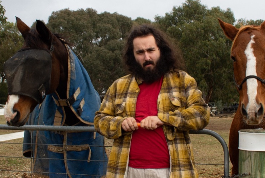 Man with beard and dark hair standing next to a horse, wearing a yellow plaid shirt.